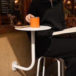 May include: An orange enamel mug sits on a small white table. The table is supported by a curved white metal leg attached to a wall. A menu board lists coffee and cheesecake prices. A person is seated, holding a book.