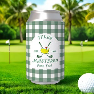 May include: A green and white checkered can cooler with the text "TYLER" and "MASTERED FORE-TEE!" on a golf course. A white golf ball sits on the green grass. Palm trees and a blue sky are in the background.