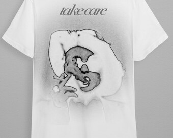 May include: White t-shirt with a black and white graphic of a person's face and the text "take care" printed on the back.