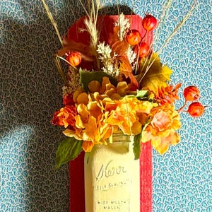 Peut inclure: Un bocal en verre avec une finition peinte en blanc et le mot "Mason" &eacute;crit dessus. Le bocal est rempli d'un bouquet de fleurs et de baies aux couleurs d'automne. Le bocal est mont&eacute; sur une plaque de bois rouge.