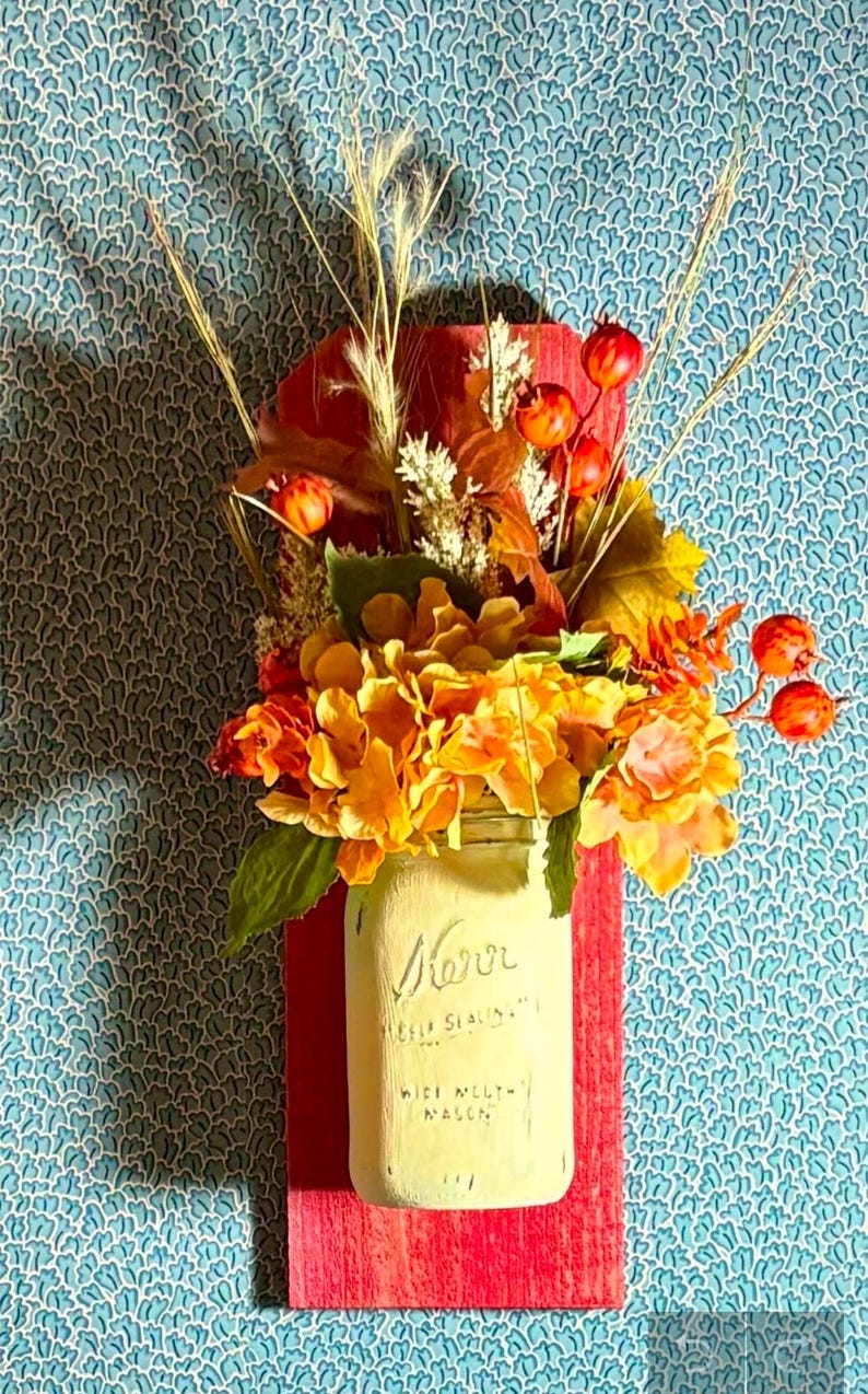 Peut inclure: Un bocal en verre avec une finition peinte en blanc et le mot "Mason" &eacute;crit dessus. Le bocal est rempli d'un bouquet de fleurs et de baies aux couleurs d'automne. Le bocal est mont&eacute; sur une plaque de bois rouge.