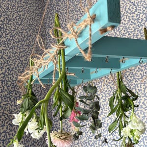 May include: A hanging floral display featuring a light blue wooden frame suspended by twine. Various flowers and greenery, including white, pink, and burgundy blooms, hang from the frame. The background is a blue and white patterned wallpaper.