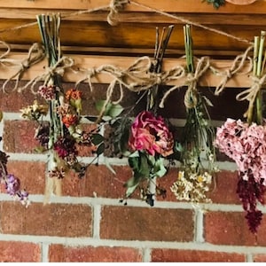 May include: A display of dried flowers and herbs hanging from a wooden beam. The bundles are tied with twine and include lavender, roses, and other greenery. The arrangement is set against a brick wall.