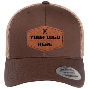 May include: Brown and tan trucker hat with a brown leather patch that says "YOUR LOGO HERE" and a "B" inside a circle. The hat has a mesh back and a snap closure.