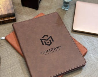 May include: A brown leather-like portfolio folder featuring a black geometric logo and the text COMPANY TAG LINE HERE printed on the front cover. The rectangular folder is layered over a lighter tan folder, showcasing a smooth, matte finish.