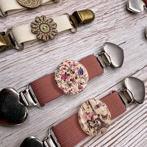 Könnte beinhalten: Dekorative Strumpfhalter-Clips mit Blumen- und Eiffelturm-Designs. Die Clips haben cremefarbene und mauvefarbene elastische Bänder, Holzknöpfe und Metallverschlüsse. Die Knöpfe weisen verschiedene Blumenmuster auf.
