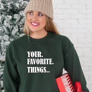 May include: A dark green sweatshirt with the text "YOUR. FAVORITE. THINGS." in white lettering. The person is wearing a beige knit beanie with a brown pom-pom.