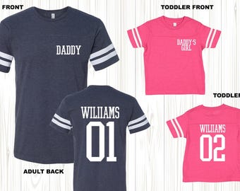 Puede incluir: Dos camisas a juego, una de adulto y otra de niño, con temática de camiseta de fútbol. La camisa de adulto es azul marino con rayas blancas en las mangas y dice "DADDY" en la parte delantera y "WILLIAMS 01" en la parte trasera. La camisa de niño es rosa con rayas blancas en las mangas y dice "DADDY'S GIRL" en la parte delantera y "WILLIAMS 02" en la parte trasera.