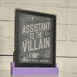 May include: A purple Kindle e-reader stand with a black Kindle e-reader displaying the cover of the book "Assistant to the Villain" by Hannah Nicole Maehrer. The book cover features a black and white illustration of a sword and a potion bottle.
