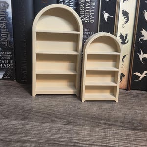 May include: Two light beige, arched, miniature wooden bookshelves with three shelves each. The taller unit stands to the left of the shorter unit, both featuring a smooth, matte finish and a curved top design, presented as standalone display pieces.