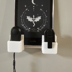 May include: White wall mount with two black controllers and a Kindle e-reader in a clear case. The Kindle screen displays a black and white image of a dragon with a crescent moon and stars. The text "kindle" is visible on the e-reader.