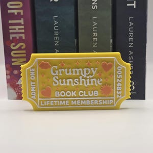 May include: A yellow ticket-shaped bookmark with the words "Grumpy Sunshine Book Club" and "Lifetime Membership" in white. The bookmark has orange heart and sun designs. The background features the spines of several books.