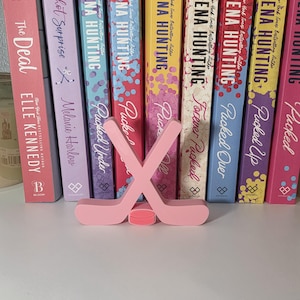 May include: A pink hockey stick and puck set in front of a row of books. The books have titles like "Pucked" and "The Deal" by various authors. The books are in various colors, including pink, blue, and yellow.