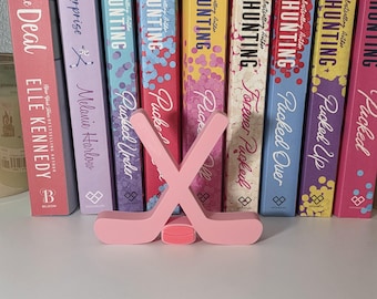 May include: A pink hockey stick and puck set in front of a row of books. The books have titles like "Pucked" and "The Deal" by various authors. The books are in various colors, including pink, blue, and yellow.