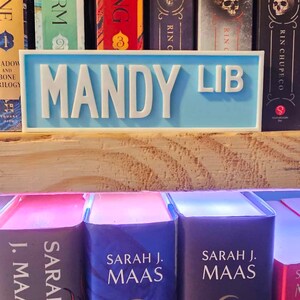 May include: A light blue bookend with the words "MANDY LIB" in white lettering. The bookend is sitting on a wooden shelf with a row of books behind it. The books have titles like "Shadow and Bone", "Siege and Storm", "Throne of Glass", and "A Court of Thorns and Roses".