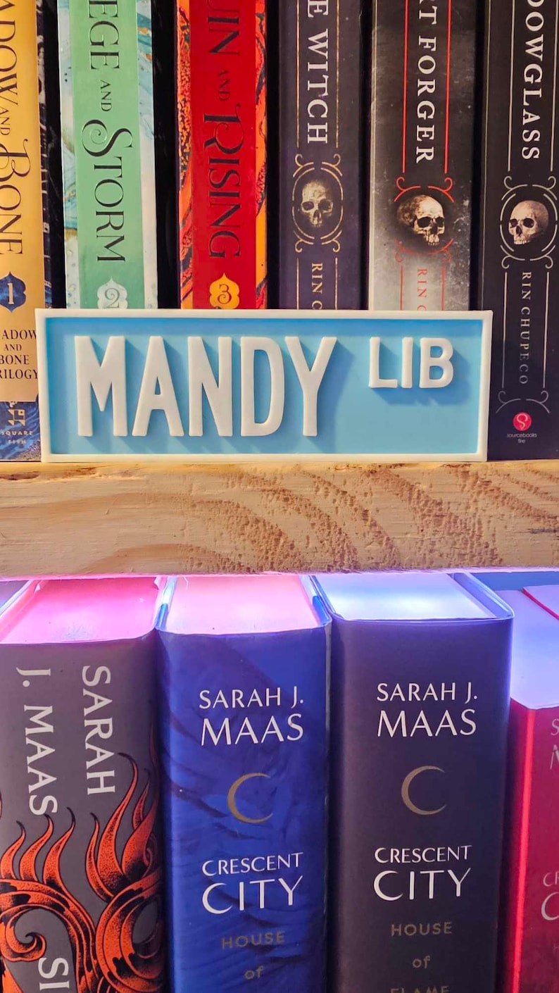 May include: A light blue bookend with the words "MANDY LIB" in white lettering. The bookend is sitting on a wooden shelf with a row of books behind it. The books have titles like "Shadow and Bone", "Siege and Storm", "Throne of Glass", and "A Court of Thorns and Roses".