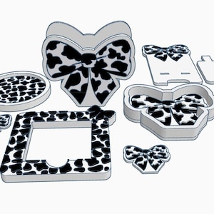 Op de afbeelding: Een verzameling van diverse 3D-geprinte objecten in een koeienprintpatroon. De items omvatten een strik, een ronde onderzetter, een fotolijst, een paperclip, een doos en diverse andere decoratieve vormen. De objecten zijn wit met zwarte vlekken.