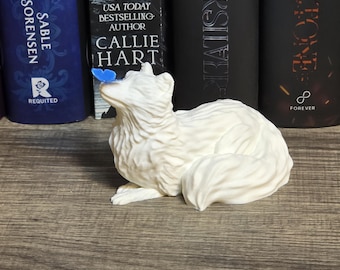 May include: A white fox figurine in a resting position with a small blue butterfly perched on its nose. The fox has detailed fur texture and is sculpted in a curled pose. The figurine is positioned in front of four books with dark spines.