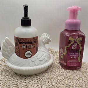 Puede incluir: Un dispensador de jab&oacute;n de cer&aacute;mica blanca con forma de gallina que contiene una botella de jab&oacute;n con aroma a hojas de oto&ntilde;o de Mrs. Meyer's Clean Day. Al lado, una botella de jab&oacute;n de manos espumoso Twisted Pepper Mint.