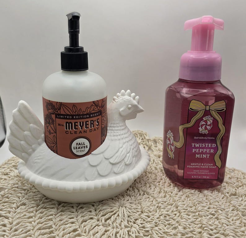 Puede incluir: Un dispensador de jab&oacute;n de cer&aacute;mica blanca con forma de gallina que contiene una botella de jab&oacute;n con aroma a hojas de oto&ntilde;o de Mrs. Meyer's Clean Day. Al lado, una botella de jab&oacute;n de manos espumoso Twisted Pepper Mint.