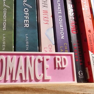 May include: A pink sign with the words "ROMANCE RD" in white lettering. The sign is on a shelf with a row of books behind it.