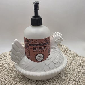 Puede incluir: Un dispensador de jab&oacute;n con forma de gallina de cer&aacute;mica blanca con una botella de jab&oacute;n Mrs. Meyer's Clean Day Fall Leaves Scent. El dispensador tiene una bomba negra y la botella de jab&oacute;n tiene una etiqueta rosa.