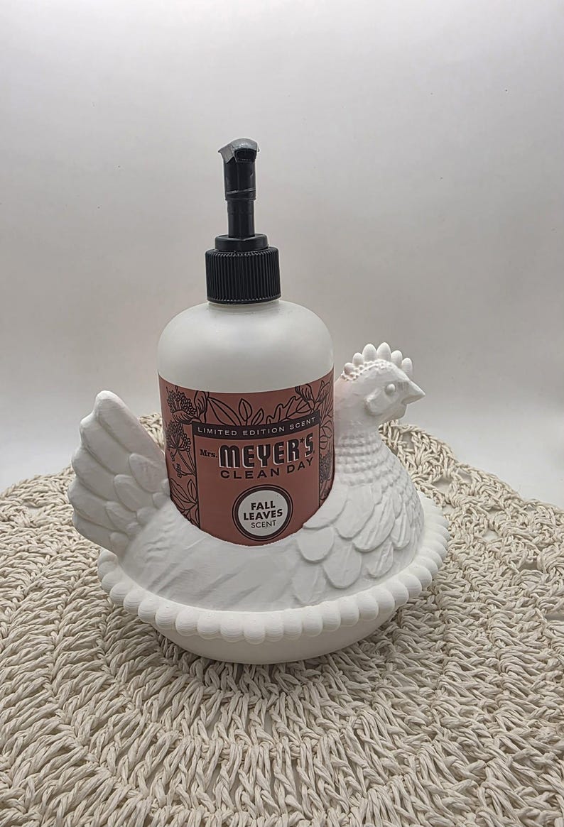 Puede incluir: Un dispensador de jab&oacute;n con forma de gallina de cer&aacute;mica blanca con una botella de jab&oacute;n Mrs. Meyer's Clean Day Fall Leaves Scent. El dispensador tiene una bomba negra y la botella de jab&oacute;n tiene una etiqueta rosa.