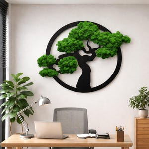 Könnte beinhalten: Schwarze Metallbaumskulptur mit leuchtend grüner Moosbepflanzung, eingebettet in einen kreisförmigen Rahmen. Das Kunstwerk ist an einer weißen Wand angebracht und erzeugt einen auffälligen Kontrast. Die Büroumgebung umfasst einen Schreibtisch, einen Laptop und eine Schreibtischlampe.