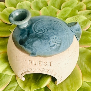 May include: A handcrafted ceramic vessel with a teal blue top and a cream-colored base. The word "GUEST" is inscribed on the base. The vessel has a unique shape and is decorated with embossed floral designs.