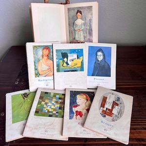 May include: A collection of vintage art books featuring various artists, including Modigliani, Picasso, and Braque. The book covers display colorful illustrations and portraits. An open book with a portrait is visible in the background.
