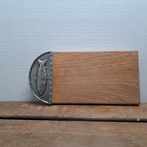 Peut inclure: Une planche à découper rectangulaire en bois avec un motif de poisson en métal décoratif à une extrémité. Le bois est brun clair et le poisson en métal est argenté. La planche est posée sur une surface en bois.