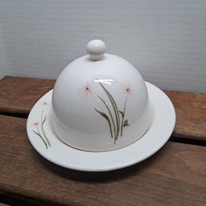 Puede incluir: Mantequera de cerámica blanca con tapa abovedada y plato a juego. El plato está decorado con un diseño floral con detalles en rosa y marrón. La tapa tiene un pequeño asa redonda.