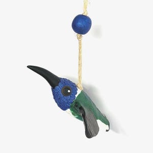 May include: A hanging hummingbird ornament featuring a textured blue head, a green body, and black wings. The bird has a long, slender black beak and a dark eye. It is suspended by a light-colored twine string that includes a single round blue bead.