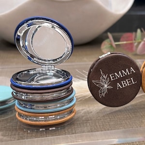 May include: Three small round compact mirrors with personalized engravings. The first mirror is brown with a butterfly design and the text "EMMA ABEL". The second mirror is light brown with the text "2 Personalized Monogram Chart". The third mirror is brown with a crescent moon design and the letter "B" in the center.