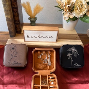 May include: Three faux leather jewellery boxes in brown, black, and tan with floral designs and personalised names. The boxes are open and show compartments for rings, earrings, and necklaces. The boxes are on a red surface with a wooden sign that reads "kindness".