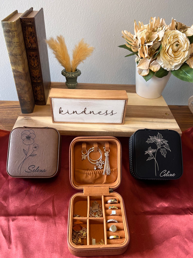 May include: Three faux leather jewellery boxes in brown, black, and tan with floral designs and personalised names. The boxes are open and show compartments for rings, earrings, and necklaces. The boxes are on a red surface with a wooden sign that reads "kindness".