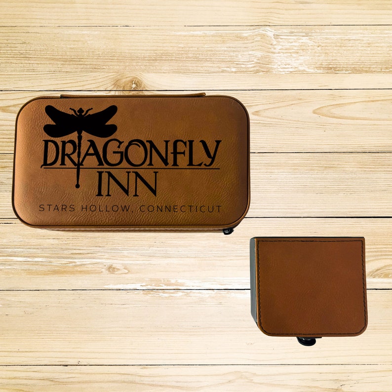 Op de afbeelding: Een bruine leren doos met een libellenontwerp en de tekst "Dragonfly Inn, Stars Hollow, Connecticut".