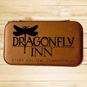 Op de afbeelding: Een bruin leren etui met de woorden "Dragonfly Inn" en "Stars Hollow Connecticut" erop gedrukt. Boven de woorden bevindt zich een zwart silhouet van een libel.