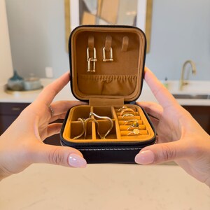 May include: A black faux leather jewellery box with a brown velvet interior. The box has multiple compartments for storing rings, earrings, and necklaces. The box is open and the compartments are filled with gold jewellery.