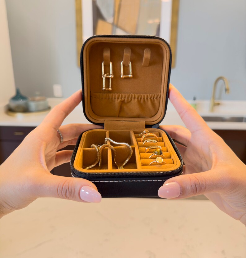 May include: A black faux leather jewellery box with a brown velvet interior. The box has multiple compartments for storing rings, earrings, and necklaces. The box is open and the compartments are filled with gold jewellery.
