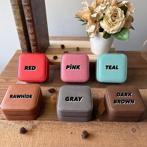 May include: Six small square jewellery boxes in different colours. The boxes are labelled with the colours: Red, Pink, Teal, Rawhide, Grey, and Dark Brown.
