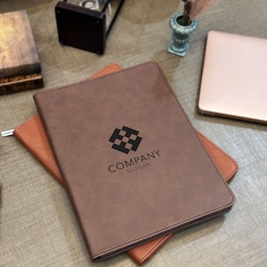 May include: A dark brown rectangular portfolio folder with a smooth texture. The front cover features a black geometric logo consisting of four squares arranged in a diamond shape, with the text COMPANY SLOGAN printed in black below the logo.