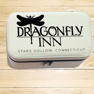 Op de afbeelding: Witte reistas voor make-up met een zwarte libellenafbeelding en de tekst "Dragonfly Inn, Stars Hollow, Connecticut".