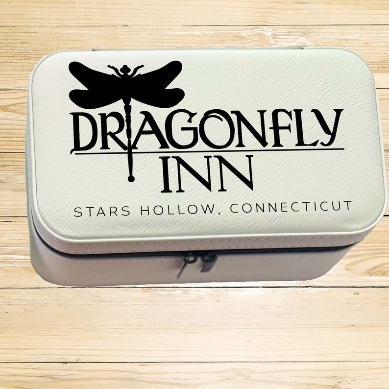 Op de afbeelding: Witte reistas voor make-up met een zwarte libellenafbeelding en de tekst "Dragonfly Inn, Stars Hollow, Connecticut".