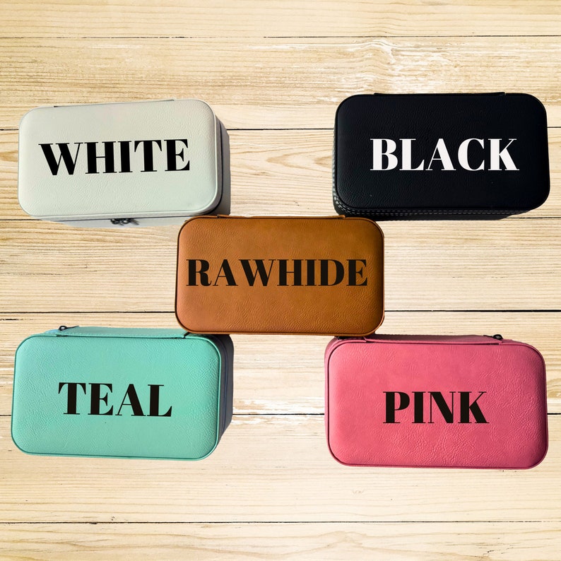Op de afbeelding: Vijf juwelendoosjes in verschillende kleuren: wit, zwart, bruin, teal en roze. Elk doosje heeft een andere kleur en een woord dat erop is gedrukt: WHITE, BLACK, RAWHIDE, TEAL, PINK.