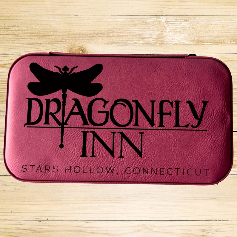 Op de afbeelding: Een bordeauxrood lederen gebonden dagboek met de tekst "Dragonfly Inn" en "Stars Hollow, Connecticut" in zwarte letters. Boven de tekst staat een libellen-silhouet.