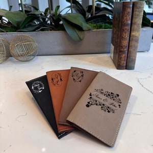May include: Four personalized passport holders in different colors and designs. The holders are made of faux leather and have a stitched edge. The designs include a floral pattern with the names "Anna & John", a monogram "A&J" in a wreath, and a monogram "A&J" in a wreath.