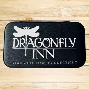 Op de afbeelding: Zwarte leren doos met een wit libellenlogo en de tekst "Dragonfly Inn" en "Stars Hollow, Connecticut".