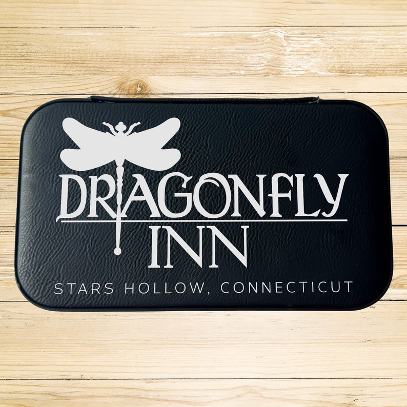 Op de afbeelding: Zwarte leren doos met een wit libellenlogo en de tekst "Dragonfly Inn" en "Stars Hollow, Connecticut".