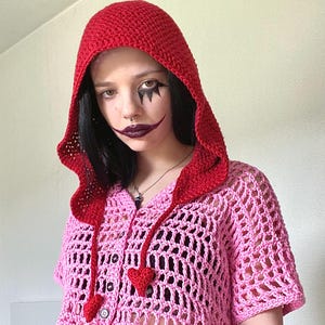 May include: A pink and red crocheted top with a hood. The top has a button closure and heart-shaped details.